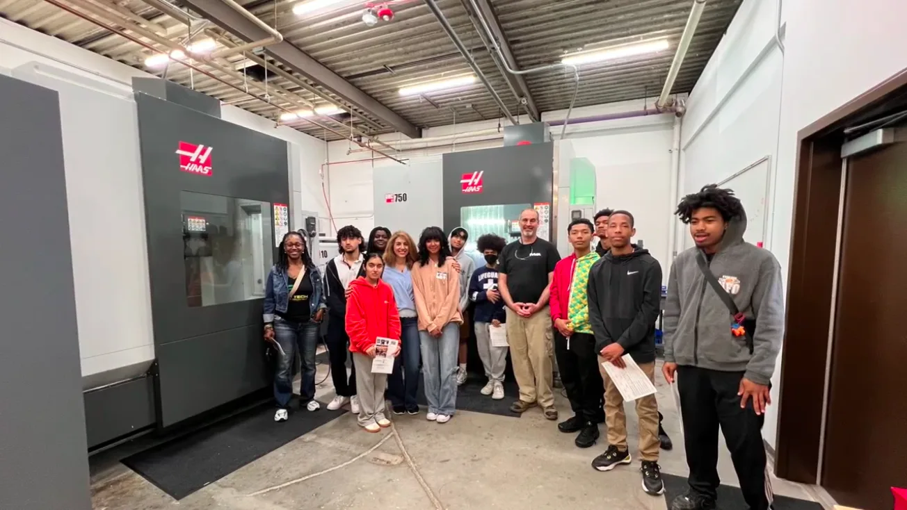 Project IGNITE students and their instructor on a tour of a manufacturing shop floor, standing beside Haas CNC machines